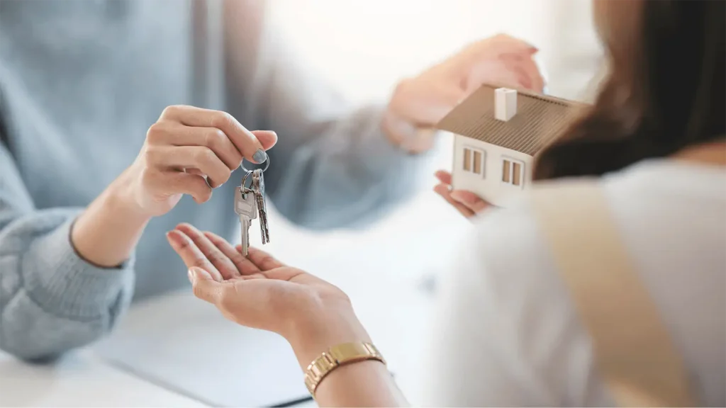 Person handing over house keys to another person holding a miniature home model, representing renting a home